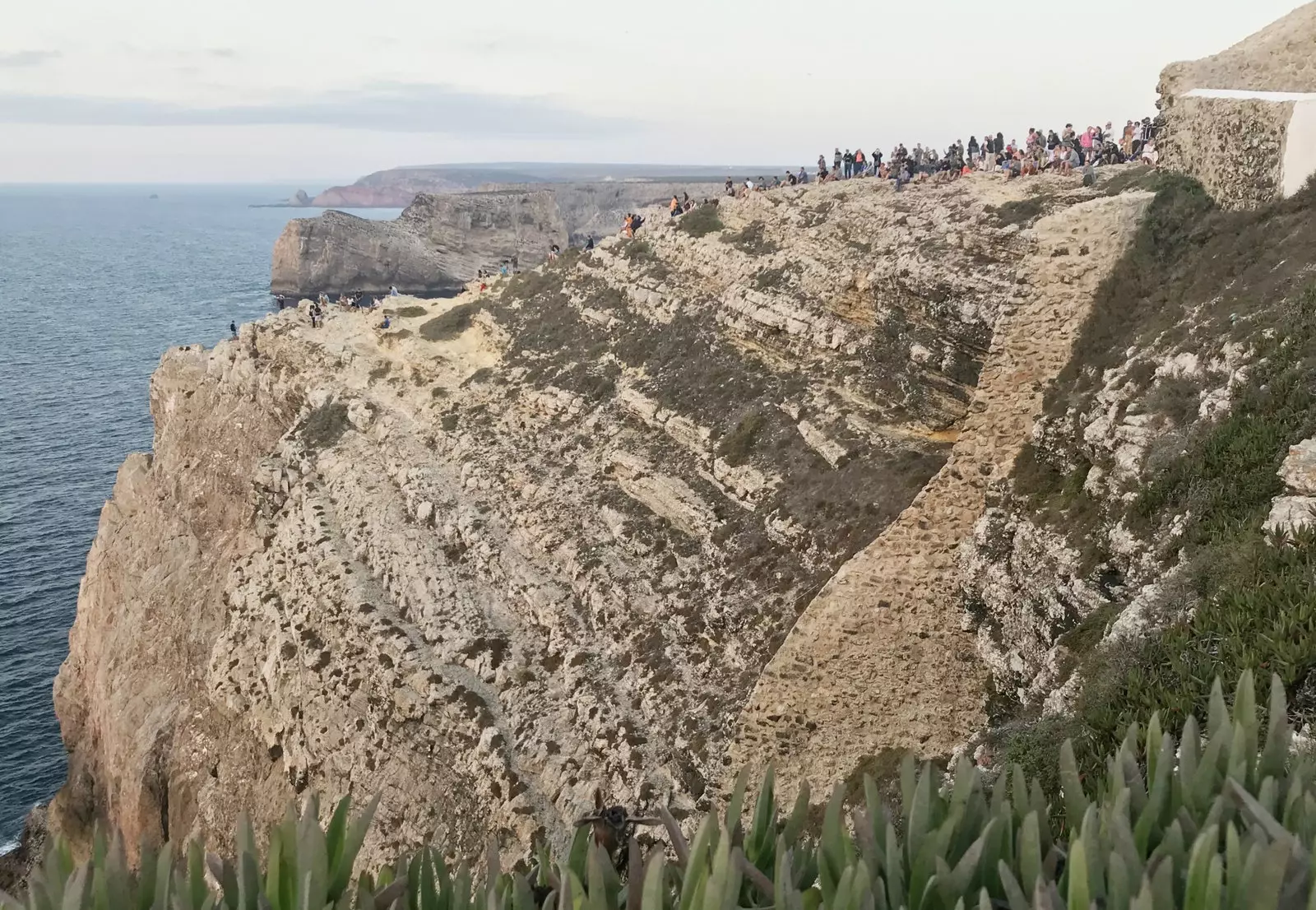 Dozens of people watching the sun disappear over the horizon on a cliff next to the Cabo de San Vicente lighthouse.