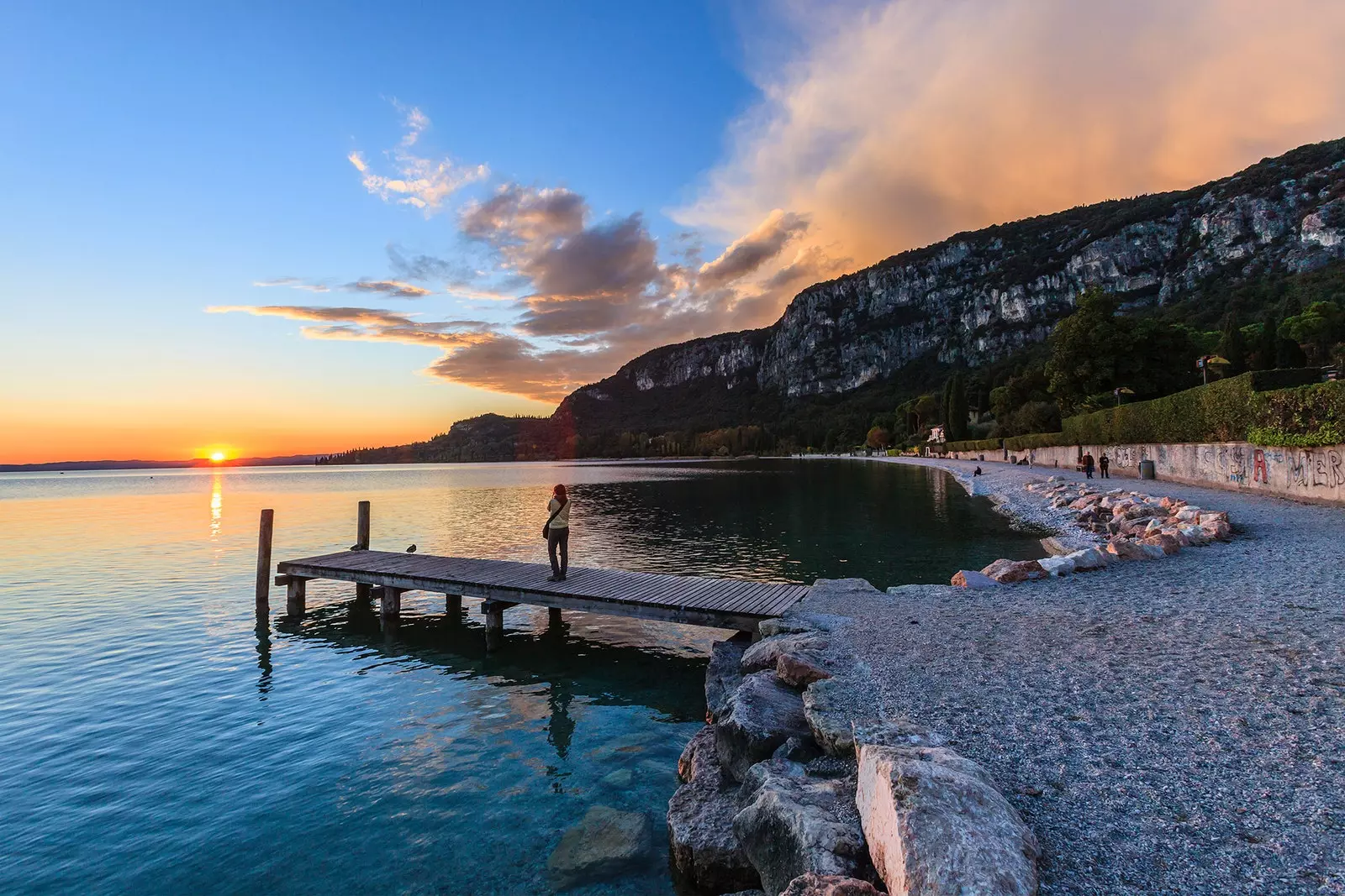 Vue op ee vun de Uferen vum Lago di Garda bei Sonnenënnergang mat Leit op der Sandstrand an op der Holzpier.