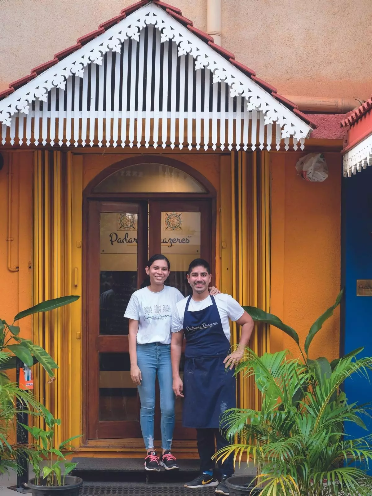 Porta com dossel de azulejo vermelho e guarnição branca com duas pessoas na frente vestidas de jeans e camiseta branca