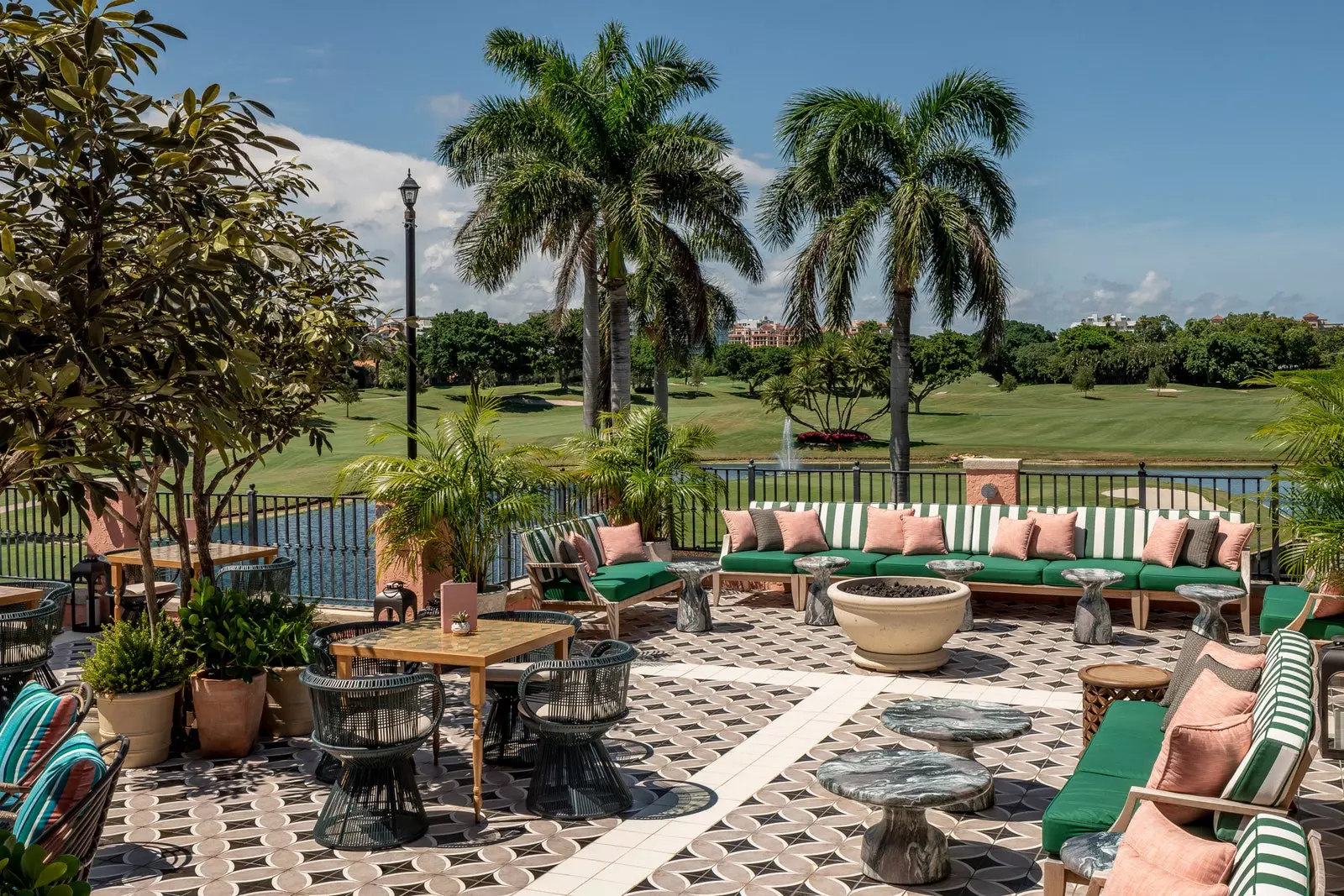 Varanda com mesas cadeiras e sofás com almofadas rosa verde e branco com vista para um campo de golfe com palmeiras