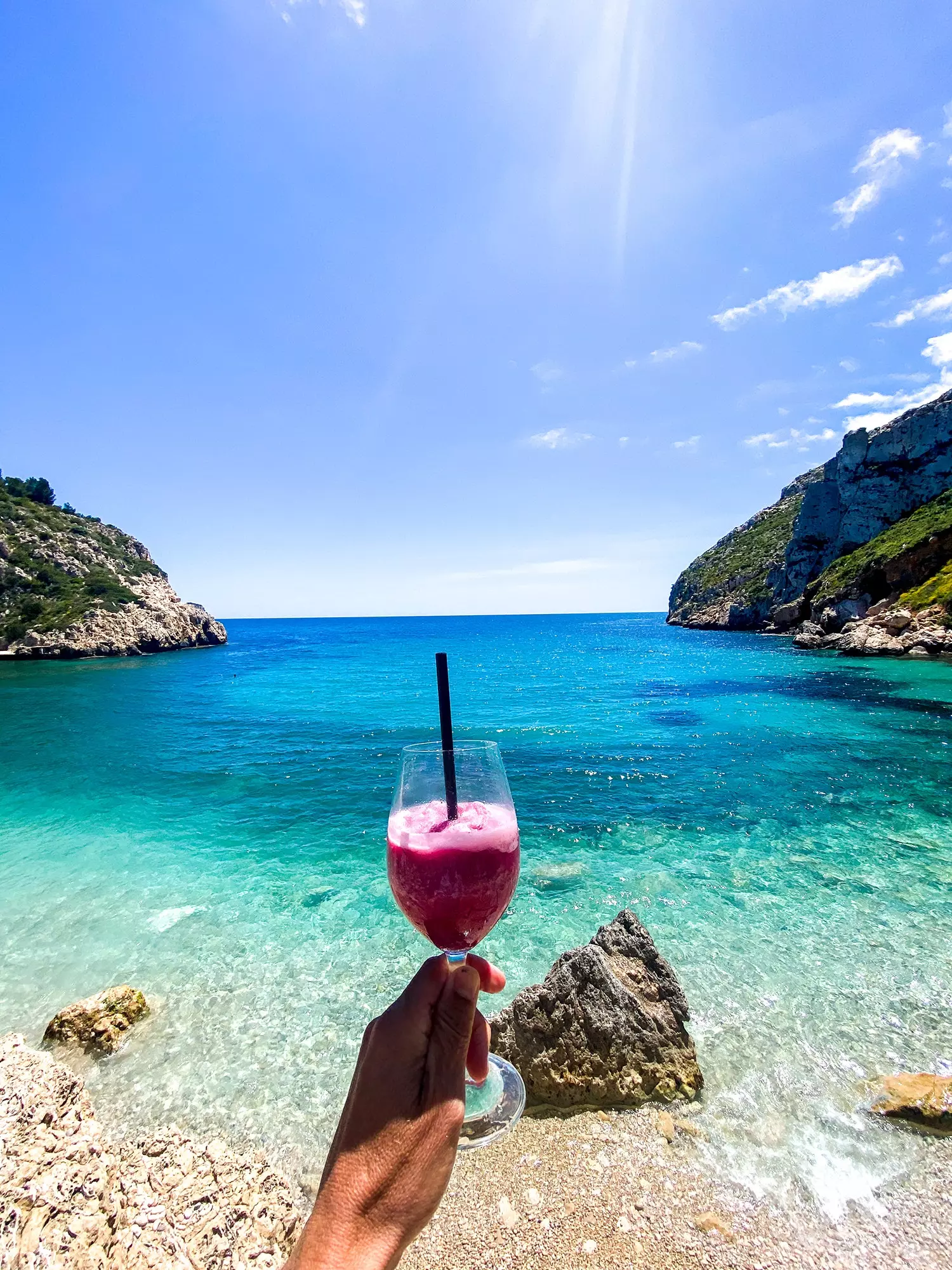 A mão segura um copo de vinho tinto de verão congelado de La Bandideta em frente ao mar azul da enseada de La Granadella.